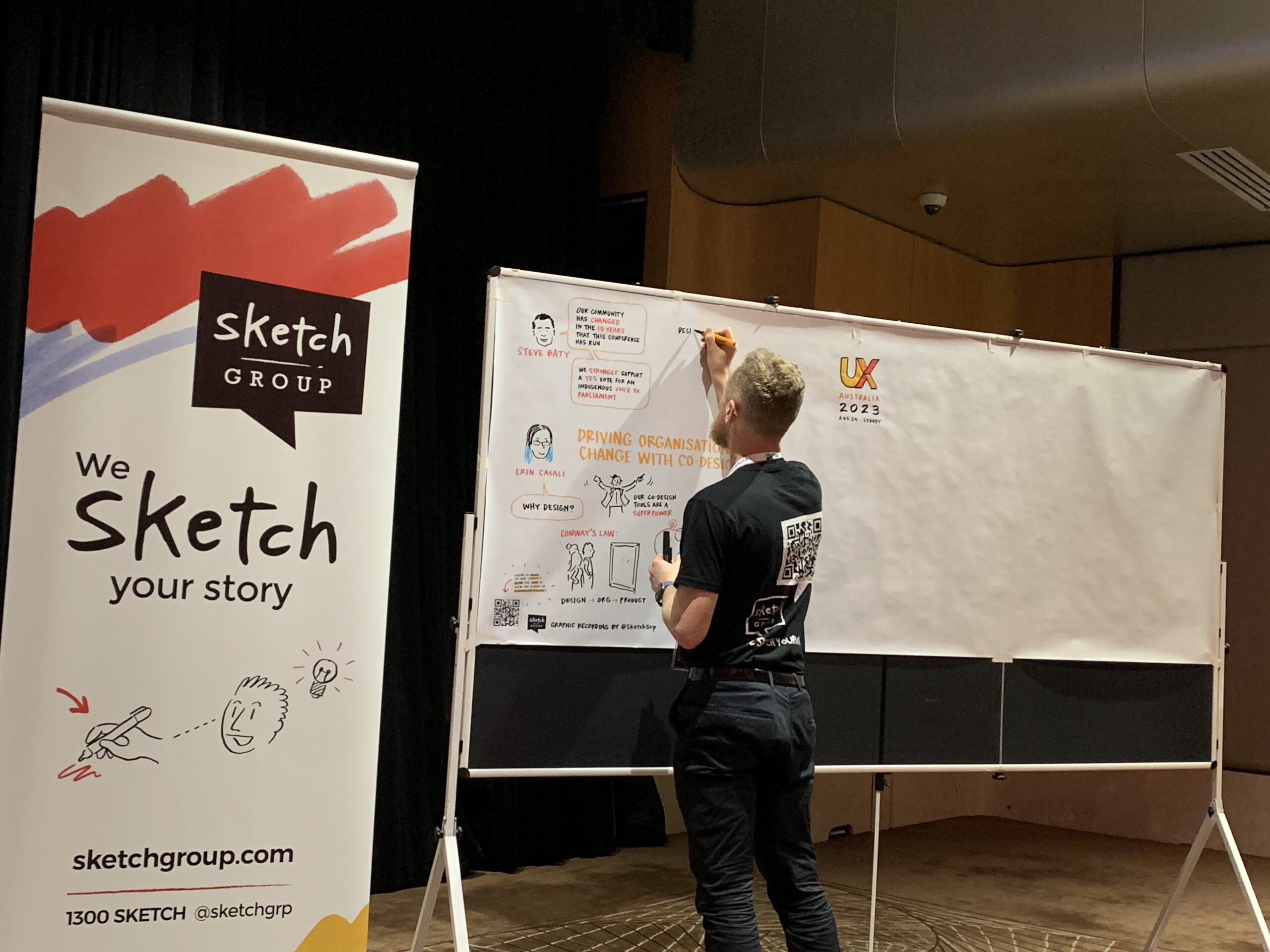 Image of graphic recorder at an event with a banner of Sketch Group next to the canvas